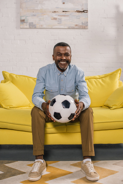smiling african american man sitting on sofa and holding soccer ball - Фото, зображення