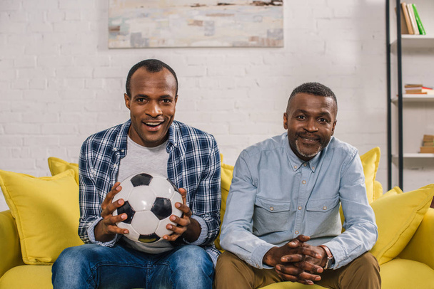 feliz afroamericano padre con adulto hijo celebración fútbol pelota y sonriendo a la cámara en casa
 - Foto, imagen