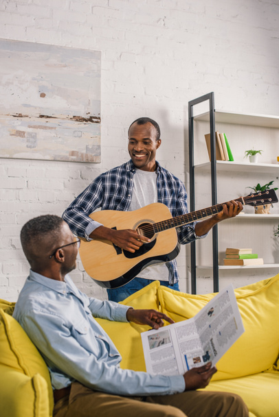 boldog magas rangú ember, újság és felnőtt fiával gitár mosolyogva egymást otthon  - Fotó, kép