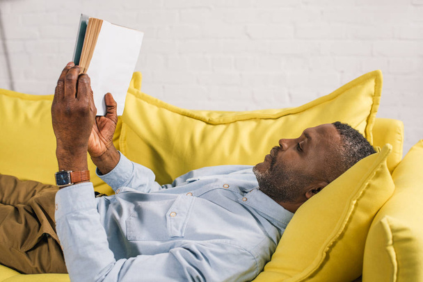 side view of senior african american man lying on couch and reading book - 写真・画像