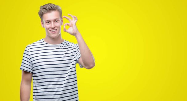 junger hübscher blonder Mann mit gestreiftem Matrosen-T-Shirt, der ok Zeichen mit Fingern macht, hervorragendes Symbol - Foto, Bild