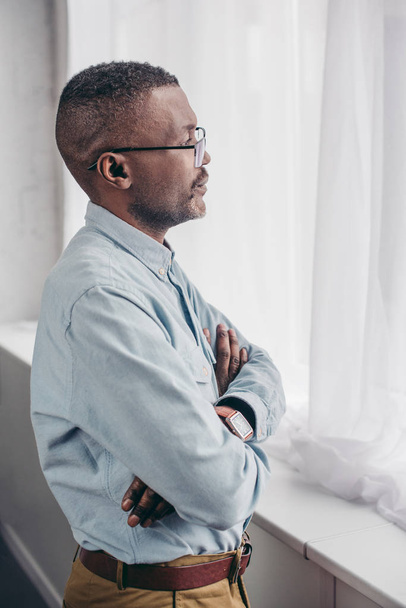 vista lateral del hombre afroamericano senior en gafas de pie con los brazos cruzados y mirando a la ventana
 - Foto, Imagen