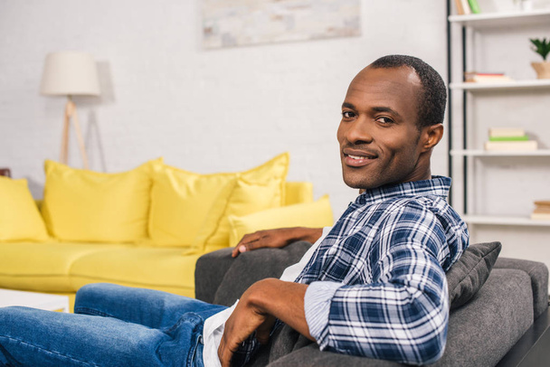 handsome young african american man sitting in couch and smiling at camera - Zdjęcie, obraz