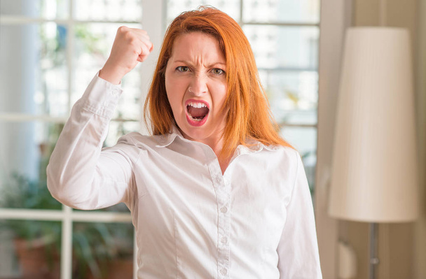 Femme rousse portant une chemise blanche à la maison ennuyée et frustrée criant de colère, folle et criant avec la main levée, concept de colère
 - Photo, image