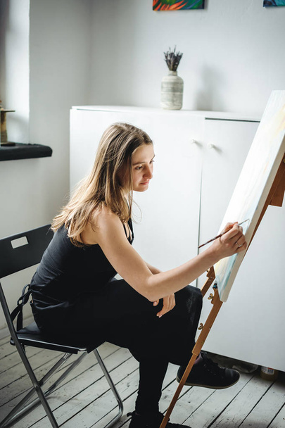 young woman in black clothes and an apron sits in a bright room and paints pictures, brushing oil paints on canvas with malbert - Fotoğraf, Görsel