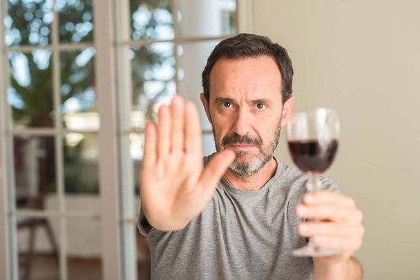 Man van middelbare leeftijd drinken van een glas wijn met open hand doen stopbord met ernstige en vol vertrouwen meningsuiting, defensie gebaar - Foto, afbeelding