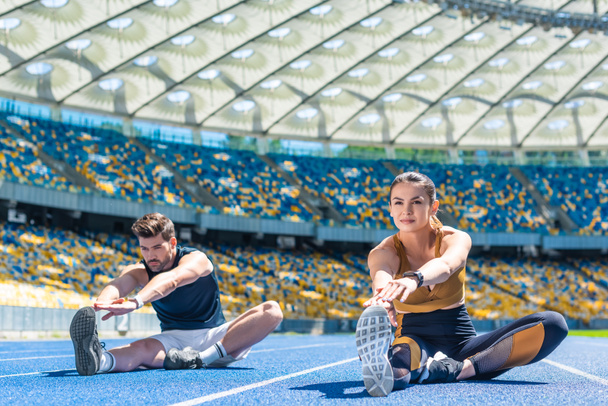 young sportive male and female joggers sitting on running track and stretching at sports stadium - Photo, Image
