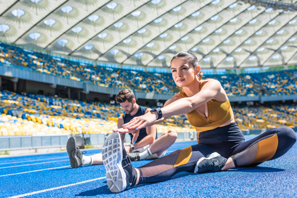 jóvenes corredores masculinos y femeninos sentados en pista de atletismo y estirándose en el estadio deportivo
 - Foto, Imagen