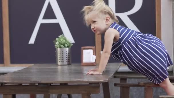 Uma menina alegre pequena está sentada a uma mesa em um café
 - Filmagem, Vídeo