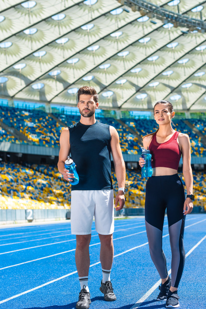 mladý sportovní pár lahvemi vody stojící na běžecké trati na sportovním stadionu - Fotografie, Obrázek