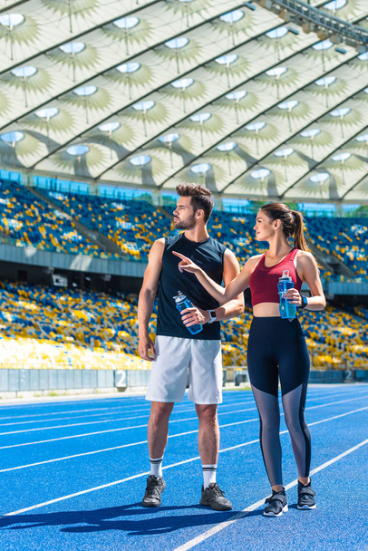 young fit couple with bottles of water chatting on running track at sports stadium - Фото, изображение