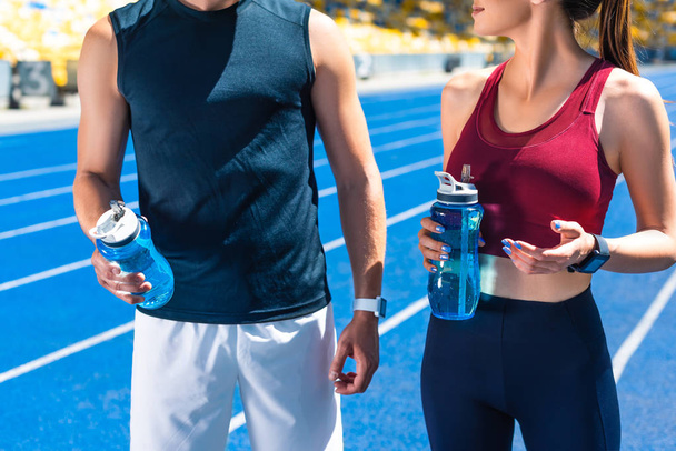 bijgesneden schot van fit paar met flessen water op atletiekbaan bij sportstadion - Foto, afbeelding