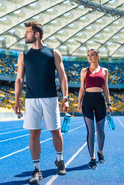 joven pareja deportiva con botellas de agua relajante en pista de atletismo en el estadio deportivo
 - Foto, Imagen
