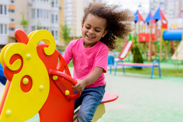smiling curly african american little child riding on rocking horse at playground  - Foto, imagen
