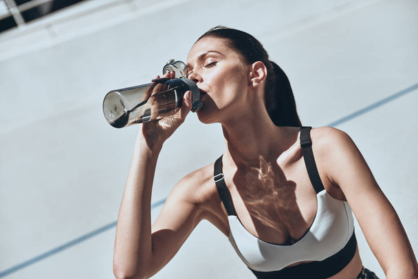 brunette woman in sports clothing resting on running track stadium and drinking water  - Photo, image