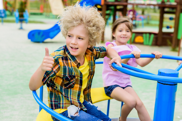 adorable curly boy doing thumb up gesture while riding on carousel with little child at playground  - Foto, Bild