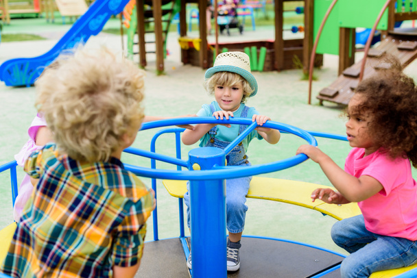 fröhliche multiethnische Gruppe kleiner Kinder auf Karussell auf Spielplatz  - Foto, Bild