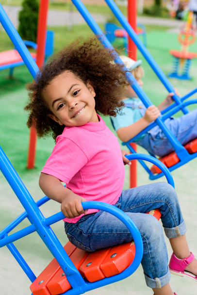 lachende krullend Afro-Amerikaanse kindje rijden op schommel in Speeltuin  - Foto, afbeelding