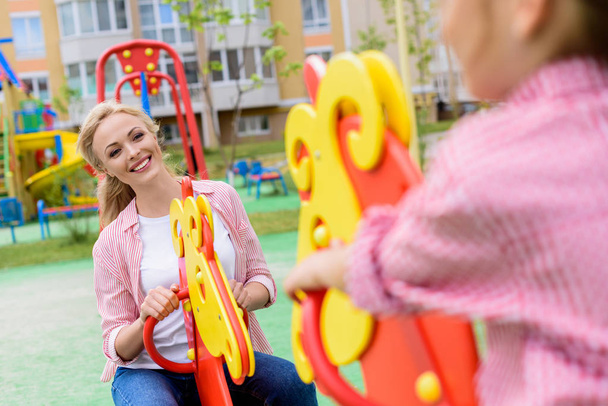 abgeschnittenes Bild eines Mädchens, das auf Schaukelpferd reitet, mit lächelnder Mutter auf Spielplatz  - Foto, Bild