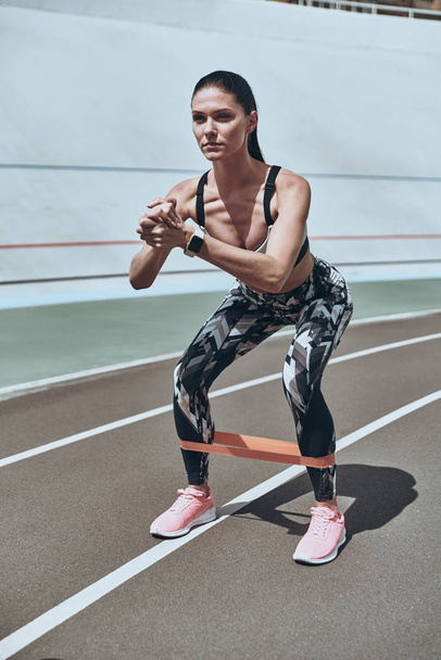 strong sportive woman crouching with resistance band on running track outdoors - Foto, afbeelding