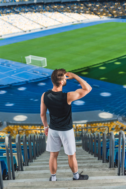 high angle view of handsome young sportsman standing on stairs at sports stadium and looking away - Photo, Image