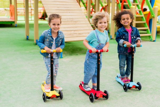 three multicultural adorable little children riding on kick scooters at playground  - Photo, Image