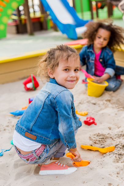 selective focus of smiling little child sitting in sandbox with plastic scoop while her friend sitting behind at playground  - Photo, Image