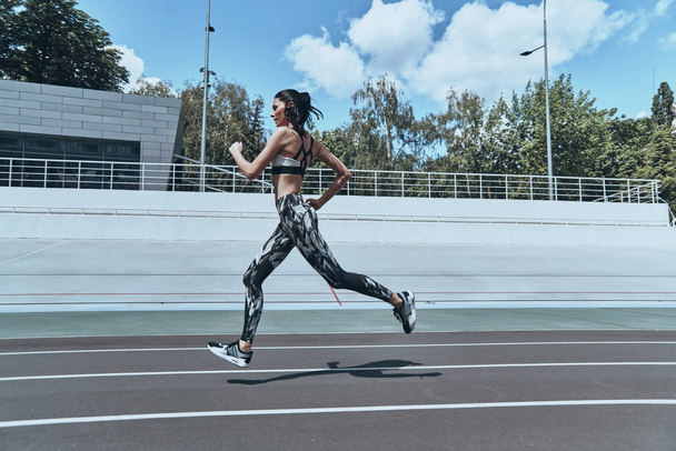 full length of brunette woman in sports clothing running on track stadium  - Foto, afbeelding