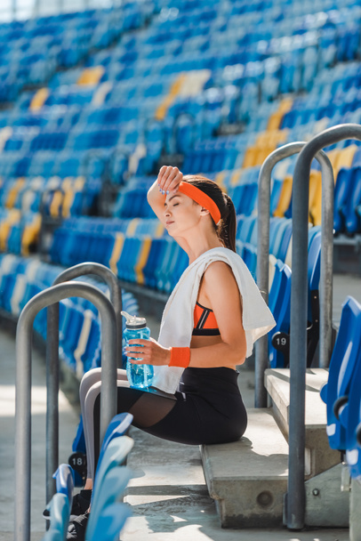 στην πλαϊνή όψη κουρασμένη νεαρή γυναίκα χαλαρώνοντας στις σκάλες και σκουπίζει τον ιδρώτα στο αθλητικό στάδιο - Φωτογραφία, εικόνα