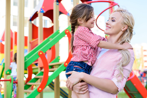 Улыбающаяся мать держит очаровательную маленькую дочь на руках на детской площадке
  - Фото, изображение