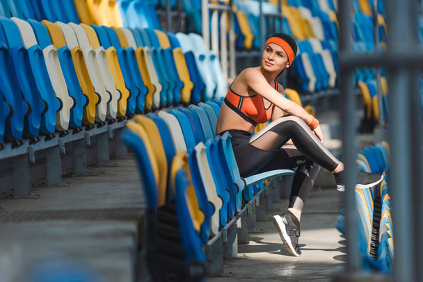 vista lateral de la hermosa joven sentada en tribunas en el estadio deportivo
 - Foto, imagen