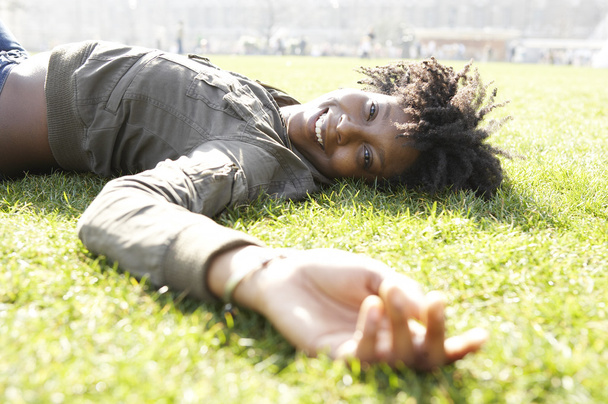 Young african american woman laying down on green grass in the city, on a sunny day. - 写真・画像