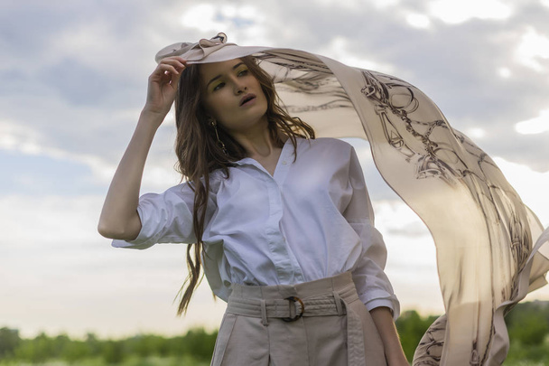 Beautiful stylish girl wearing a trendy white shirt, beige trousers and a pareo poses outdoors against a background of meadow full of wild flowers and blue cloudy sky. Advertising design. Copy space. - Φωτογραφία, εικόνα