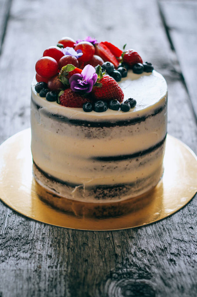 Chocolate cake with creamy cream decorated with strawberries, blueberries, grapes and violets on a wooden background - Photo, Image