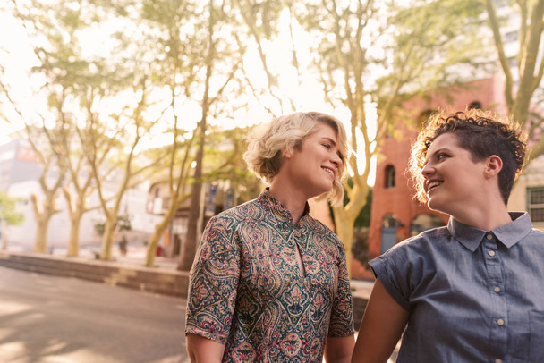 Affectionate young lesbian couple smiling and walking hand in hand together along a city street on a sunny day - 写真・画像