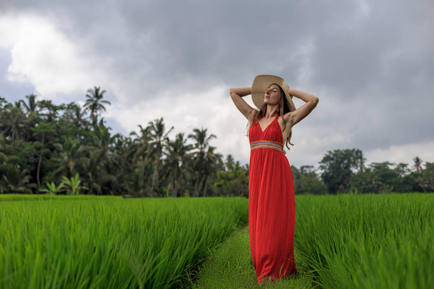 Piękna młoda kobieta w czerwonej sukience dotyka słomkowego kapelusza. Dziewczyna chodzić na typowym azjatyckim wzgórzu z uprawy ryżu, górski kształt zielony kaskadowy ryż pola tarasy paddies. Ubud, Bali, Indonezja. - Zdjęcie, obraz