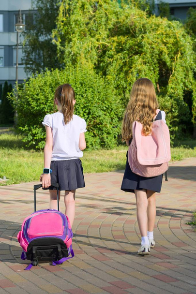 Rear view of two schoolgirl girlfriends elementary school students walking with school bag in the yard. - 写真・画像