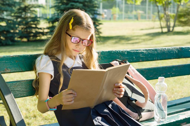 Portrait of schoolgirl primary school with backpack on a bench reading book, background school yard. - Foto, immagini