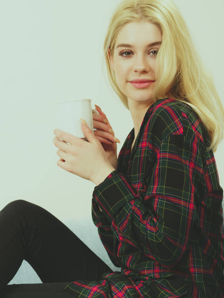 Pretty hipster young adult teenage woman sitting on sofa enjoying her leisure time drinking coffee or tea from mug. - 写真・画像