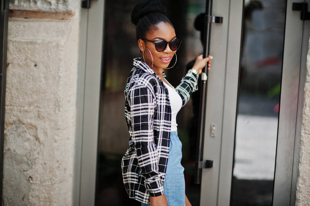 Hip hop african american girl on sunglasses and jeans shorts. Casual street fashion portrait of black woman. - 写真・画像