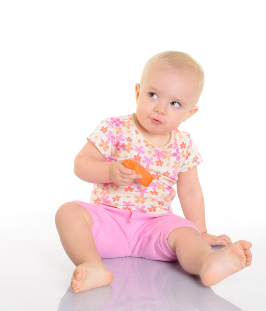 süßes kleines Baby isst eine Möhre auf dem Boden sitzend auf weiß - Foto, Bild