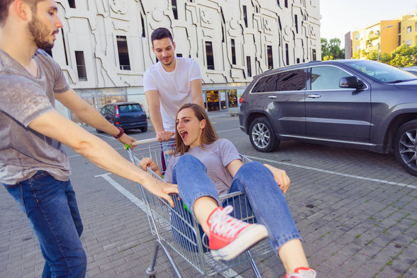 Two guys driving shopping trolley with girl in it on a parking lot - Foto, Imagem