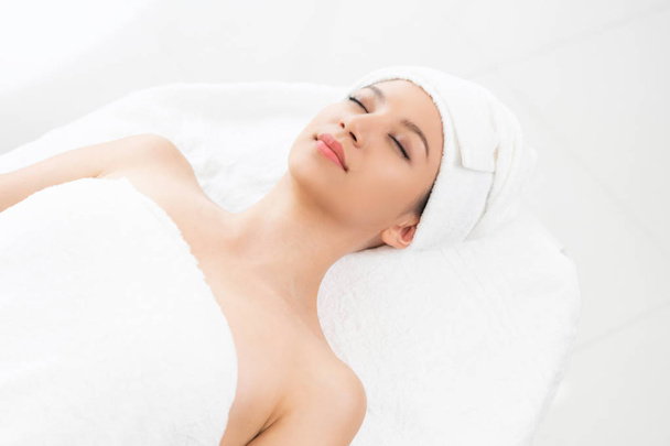 Relaxed young woman lying on spa bed prepared for facial treatment and massage in luxury spa resort. Wellness, stress relief and rejuvenation concept. - Fotó, kép