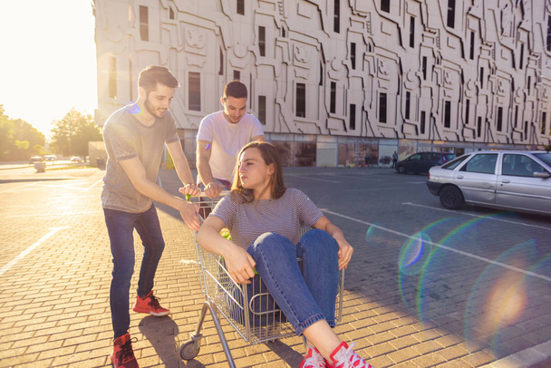Teenage girl in shopping trolley having fun while two guys is pushing her - Photo, Image