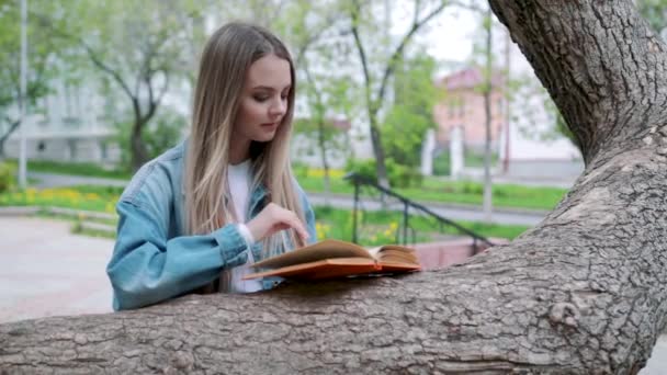 Beautiful serious girl in jeans jacket and glasses reads book, against summer green park - Footage, Video