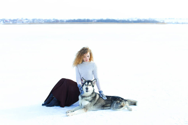 Белая девушка в свитере, коричневой юбке и варежках, сидящая на снегу и обнимающая собаку Хаски
. - Фото, изображение