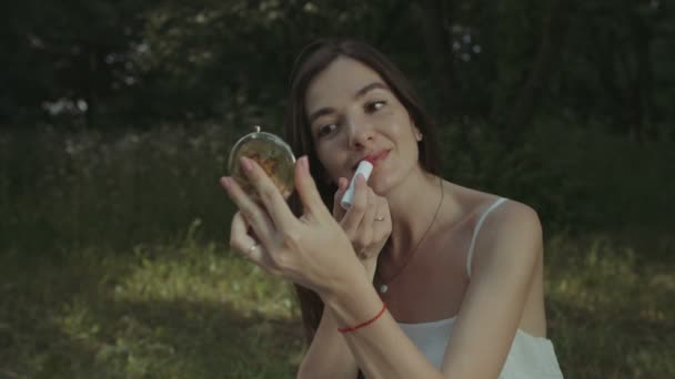 Charming woman applying lipstick in park - Filmati, video