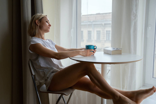 Молодая женщина с чашкой кофе читает книгу сидя дома расслабляясь у окна
 - Фото, изображение