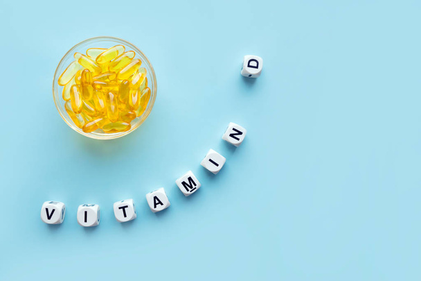Keltaiset kapselit pyöreässä lasikulhossa ja sana D-vitamiini valkoisista kuutioista, joissa kirjaimet sinisellä pohjalla. VITAMIINI D tarkoittaa tervettä ja lääketieteellistä konseptia. Auringonpaiste vitamiini terveyshyötyjä
 - Valokuva, kuva