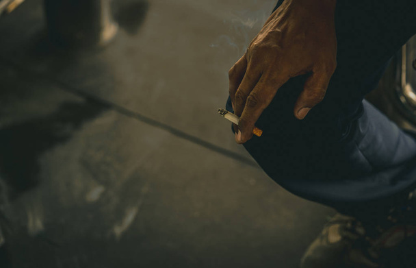 Азиат с грязной рукой держит сигарету и курит. Бедные азиаты сидят и курят сигареты во время перерыва или после тяжелой работы. Никотиновая зависимость. Канцерогены вызывают рак легких
.  - Фото, изображение
